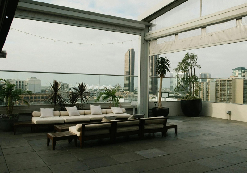Outdoor rooftop lounge area with modern seating, potted plants, and city buildings visible in the background.
