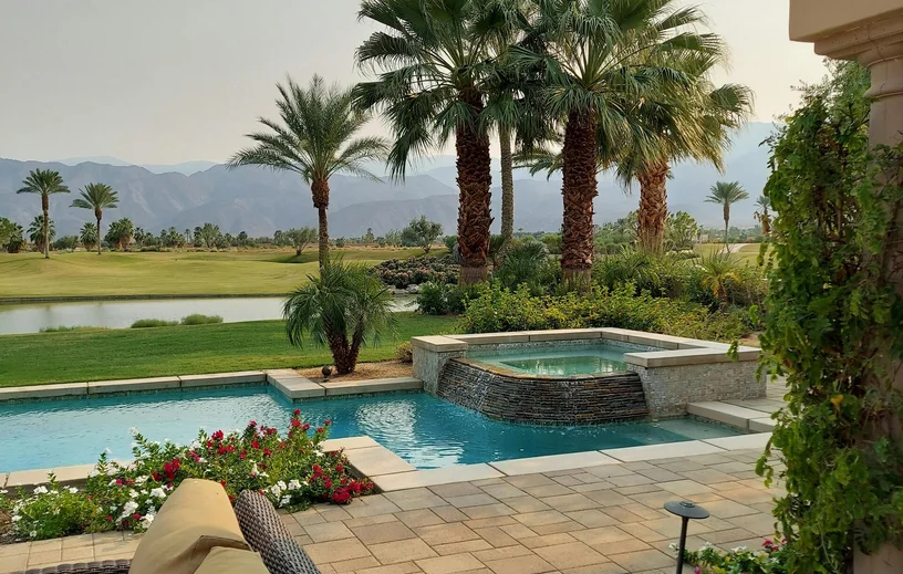 A backyard with a pool, hot tub, palm trees, landscaped garden, and a golf course in the background.