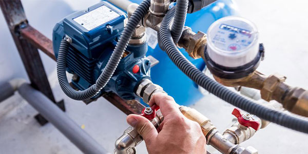 A hand adjusts a valve on a water pump system with pipes, a pressure gauge, and a blue tank in view.