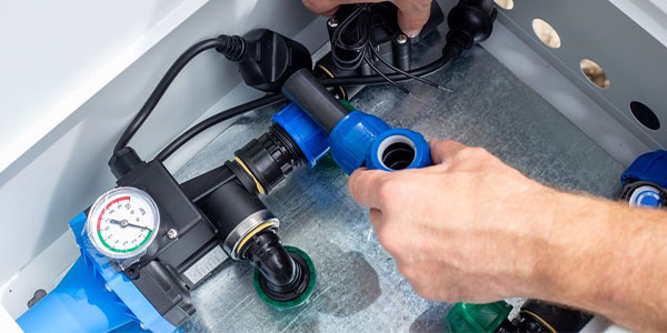 Hands adjusting blue and black plumbing fittings near a pressure gauge inside a metal enclosure.