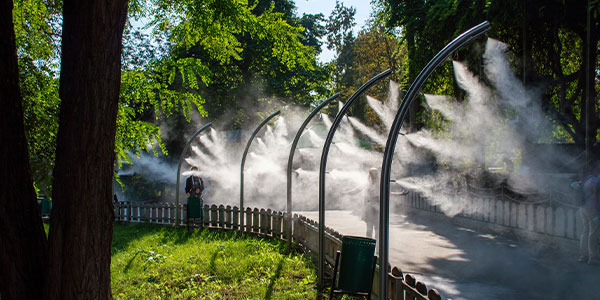 Misting arches spray fine water mist over a pathway in a shaded, tree-lined park area with a fence and trash bins.