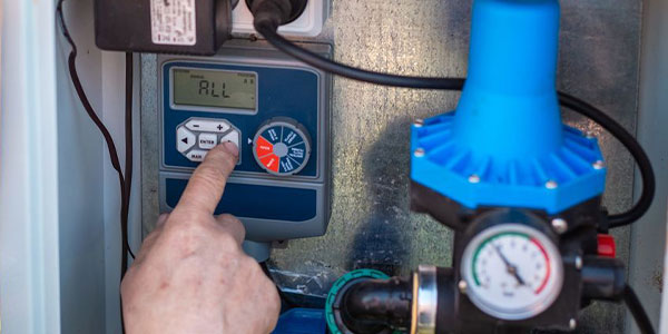 A hand adjusts settings on an electronic control panel next to a pressure gauge and blue valve apparatus.